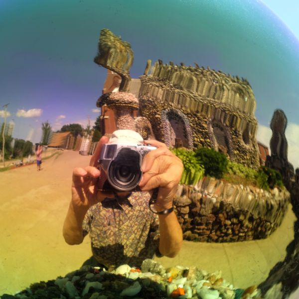 Self-portrait with a gazing ball at the Dickeyville Grotto. Richard Quinney is standing with his camera obscuring the view of his face. Shrines made of stone, mortar, and colorful rocks and objects are behind him. Bushes, trees, and decorative plants are growing along the walkway near to the shrine. Another man in the distance is taking a photograph of the shrine.
