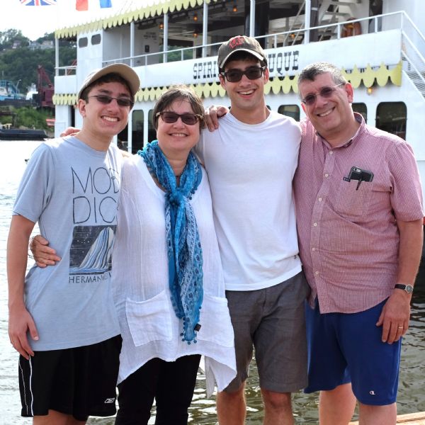 Group portrait of three men and a woman, all wearing sunglasses. The two younger men are wearing baseball hats, and the woman is wearing a blue scarf. Behind the group is a river cruise ship sitting docked in the Mississippi river, flying both British and French flags.