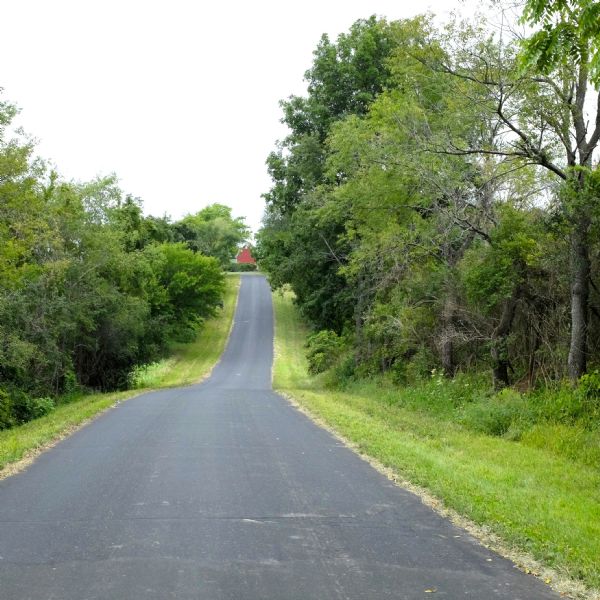 View of a long, paved driveway, bordered by trees and bushes, that goes down and up a hill. A red barn is partially visible among trees at the end of the driveway.