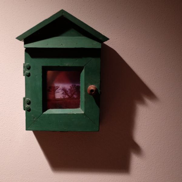 Close-up of green box in the shape of a house hanging on a pink stucco wall. A pink photograph of trees is framed by the hinged door of the box.