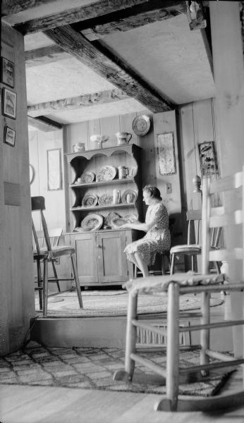 View through room towards Leonore Middleton, in another room, holding a majolica plate. She is sitting next to an open dish dresser which holds other pottery pieces. The wood paneled room has an exposed beam ceiling and wood floors.  