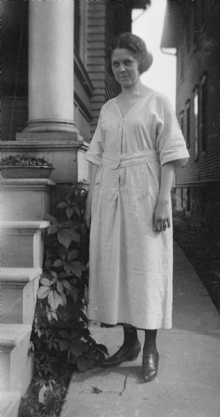 Leonore Middleton posing standing next to a pan of freshly picked strawberries, which are resting on a porch step next to a wooden column on the porch. She is wearing a wedding ring. The narrow sidewalk where Leonore is standing runs between two closely spaced houses.