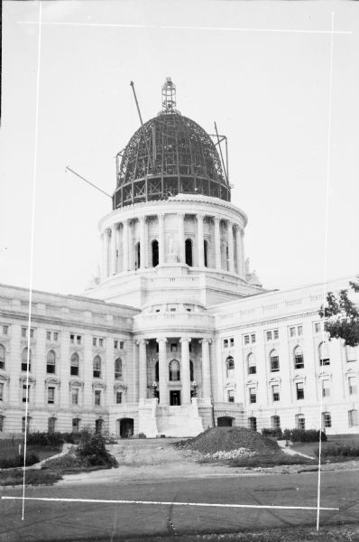 View across street towards temporary derricks protruding from the steel framework of the dome on the Wisconsin State Capitol. The lantern cage tops the dome.  The colonnade and some of the statuary at the base of the dome are in place. There is a pile of dirt in front of the stairs.