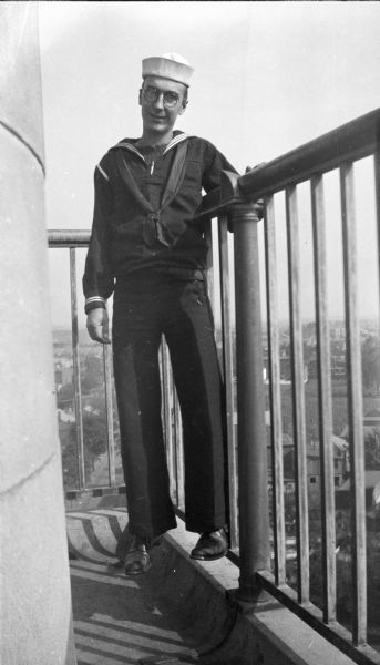 Forest Middleton, in his U.S. Navy dress blues uniform, posing and standing on a metal railing on the platform of a tall structure, possibly a lighthouse or water tower. There are buildings below in the background.