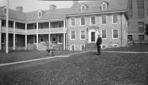 Forest Middleton, in his U.S. Navy dress blues uniform, posing on the lawn of the 1758 Old Barracks in Trenton, New Jersey. On the right is a two and one half story stone house, which shares a wall with the ell shaped barracks building, also two and one half stories, which has columns supporting a second story gallery. There is a cannon resting on a stone platform on the front lawn.  