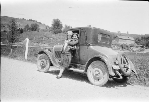 Forest Middleton, sitting in the driver's seat, embracing his wife Leonore who is standing outside the car, leaning against the door of the 1927 Chevrolet coupe. The car is parked at the edge of a gravel country road and there is a barn in the background. The car has a spare tire mounted behind the rumble seat but is missing its rear bumper.