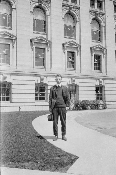 An unidentified young man standing on a curved sidewalk in front of the Wisconsin State Capitol. He is holding his hat in his right hand and is wearing a suit and tie, with a heavy sweater under his jacket. There are flag shaped cut-outs in nearly every window visible.