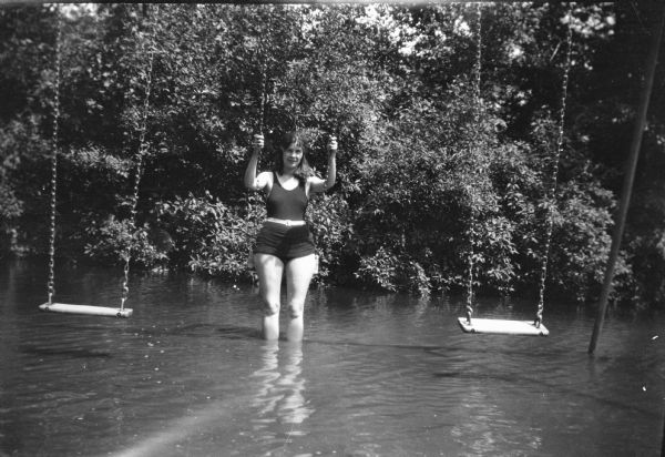 An unidentified woman standing in shallow water holding the chains of a swing. There is an unoccupied swing on either side of her, and a support post is on the right. Shrubs are on the shoreline behind her.