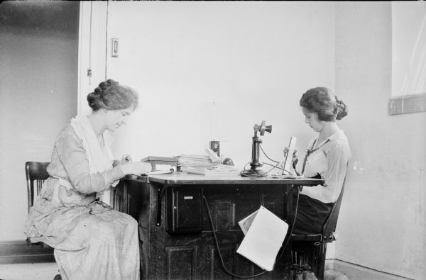 Leonore Middleton, right, and an unidentified woman sitting on opposite sides of a wooden desk. Leonore is holding a slide rule, and the other woman is looking at a small pad of paper. There is a telephone on the desk and a small box labeled "Household [?] Account Case."  A telephone book or city directory is hanging on the side of the desk, and "3 F Laundry" is visible on the cover. Leonore has a ribbon pinned to her blouse.