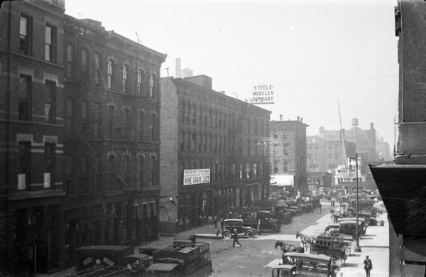 An elevated view of Dearborn Street businesses, showing trucks and horse-drawn wagons parked in front of a row of mainly four-story brick warehouse buildings. There are pedestrians on the street and sidewalks. A rooftop sign advertises the Steele-Wedeles Company, a wholesale grocery distributor located at 312-322 North Dearborn Street. The firm was in business from 1862 to 1953. A sign on the front of another building advertises "Forbidden Fruit, 'Frutta Proibita,' Wine Grape Juice."