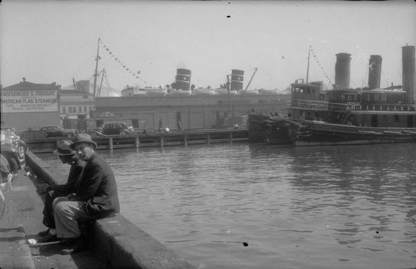 The funnels (smokestacks) of two large steamships are visible over warehouse buildings on a pier of a southern American port. Both ships have nautical flags flying and lifeboats are seen over the roof of the building. The ship on the left has a light-colored star painted on each funnel. Three tugboats are moored on the near side of the pier. Two men wearing suits and hats are sitting on a low wall in the foreground, left. One of the men, who is smoking a pipe, is looking over his shoulder at the photographer. There are cars parked along the pier. A sign painted on the building at left advertises: "Passenger & Freight Service on American Flag Steamers. Cuba — Dominican Republic  Mexico — Puerto Rico."