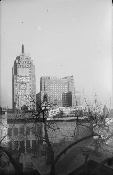 Mariner Tower, built in 1930 at 606 West Wisconsin Avenue, standing to the left of the 1927 Schroeder Hotel in this elevated view looking east over lower buildings and large trees. The clocktower of Union Train Station is on the right. Lighted signage on the Mariner Tower reads: "Save at The Boston Store" and "It's Time for Graf's, The Best What Gives[sic]" and also "Drink Hydrox Cola." There is a billboard in the foreground advertising Calvert whiskey.