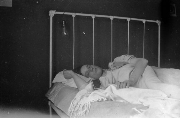 A young man, possibly Forest Middleton, lying on a painted iron bed with a bed spread covering him. He is holding a lighted cigarette in his left hand; a cigarette box or pack is lying on his right arm. There is a bare light bulb at the head of the bed, with the cord wrapped around the frame of the headboard.