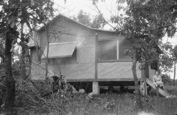 Leonore Middleton sitting on wooden steps leading to the screened porch of a tar paper shack. There is an awning over the side window and a short brick chimney supported by a wooden frame. A swimming suit is draped over a tree branch near the porch. Leonore is holding a carpenter's saw in her right hand. There is a pile of brush on the ground on the left.