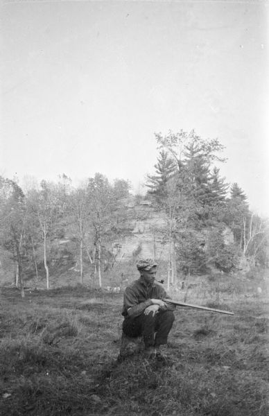 An unidentified young man sitting on a low stump with a shotgun resting in the crook of his left arm. He is wearing a plaid cap and smoking a pipe. There is a tall rock outcropping behind him, and most of the trees have lost their leaves.