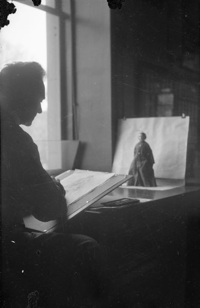 An unidentified man is seen in profile near a window. He is sitting at a table sketching a mannequin or doll which is standing against a plain, light-colored paper background. There is a small watercolor palette on the table.  