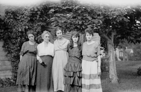 Cora Robbins Judkins, second from left, posing with her four daughters standing beside a house. The daughters are, from left, Leonore Judkins Middleton, Bonnie, Juanita and Blanche. There is a vine on the wall of the house and a catalpa tree on the right. There are houses in the background.