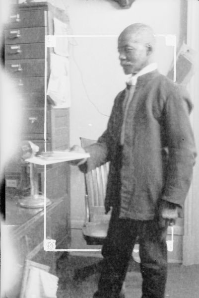 An unidentified Japanese man standing in front of a desk holding a stack of letters in his right hand. He is wearing a ring on his left hand. There is a telephone on the desk and a stack of file drawers against the wall.