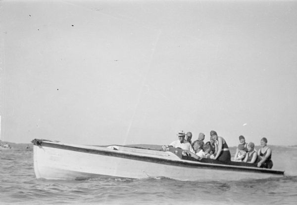 An unidentified young man, smiling broadly, is driving a painted wooden boat with inboard motor. There are nine women passengers. The women are wearing bathing suits and caps. An older man with hat and tie is riding in front, on the captain's right.  