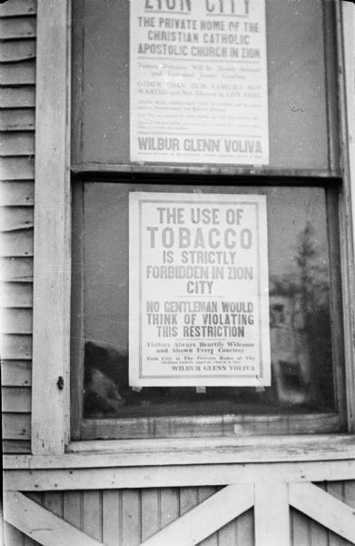 Two signs are taped to the inside of the glass in a double hung window in a wood sided building. The photographer is reflected in the lower left corner of the window. The upper sign identifies Zion City as "The Private Home of the Christian Catholic Apostolic Church in Zion." The lower sign states: "The Use of Tobacco Is Strictly Forbidden in Zion City." Both signs carry the name of Wilbur Glenn Voliva, the head of the Christian Cathlic Apostolic Church in Zion. Voliva was the successor of faith healer John Alexander Zowie (1847-1907) who founded Zion City in 1900 and was its leader until his death. Zion City was essentially a theocracy until "independents" deposed Voliva in 1937.  