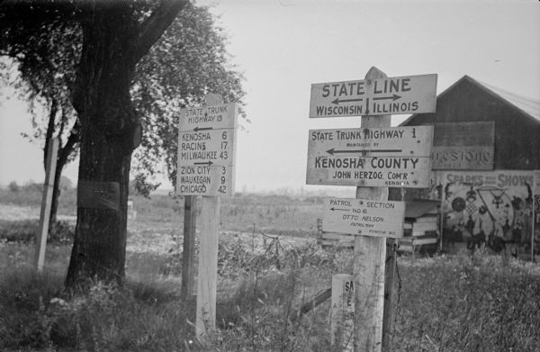 A sign post next to a survey marker, lower right, marks the state line between Wisconsin and Illinois south of Kenosha. Signage on that post identifies State Trunk Highway 1; the sign on the left identifies State Trunk Highway 15 and the directions and distances to towns in Wisconsin and Illinois. There are also signs on the barn in the background for Firestone Tires and for Sparks World Famous Shows. 