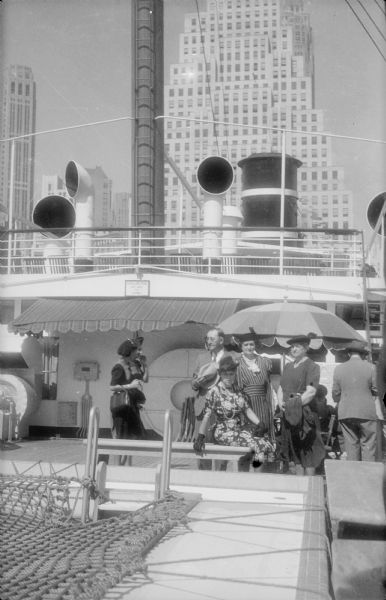 Well-dressed men and women have gathered on the deck of a cruise ship. The swimming pool in the foreground is covered with rope netting. Four dorade boxes (ventilators) and a large funnel (smokestack) are on the upper deck, as well as a mast with its ladder. The skyscraper at 120 Wall Street dominates the background.