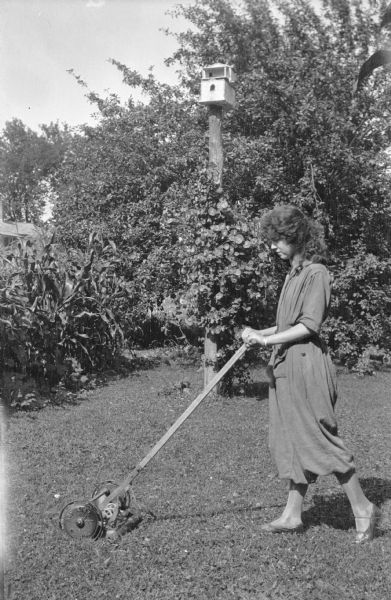Juanita Judkins pushing a "Run Easy" reel lawn mower. She is wearing heavy fabric coveralls with wide legs which end at mid-calf, exposing silk stockings. There is corn growing on the left. In the background, an ornamental vine climbs a post which is topped by a birdhouse.