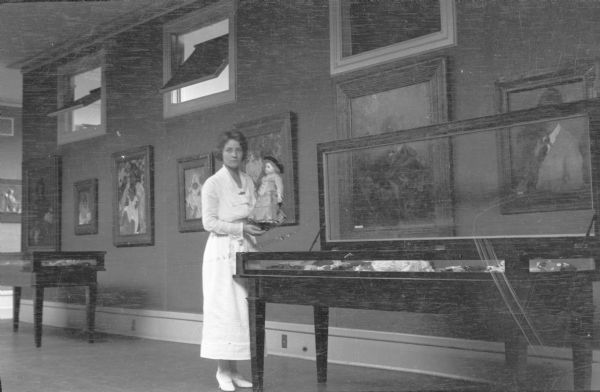 An unidentified woman is holding a doll while standing beside an open glass topped display case. She is in a gallery or museum, with paintings hanging on the wall. Two transom windows in the wall behind her are tilted open.