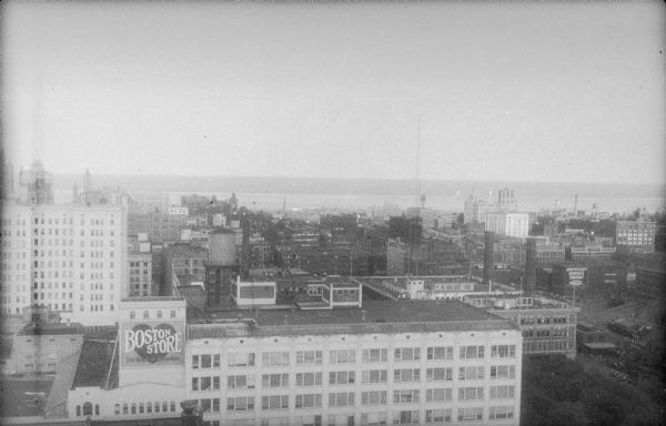 An elevated view of downtown Milwaukee, with Lake Michigan in the background. The eight story 1920 addition to the Boston Store is in the foreground, identified by a large sign reading: "Boston Store The Heart of Milwaukee." There is a water tower on the roof. The tall building on the left is the Majestic Theater Building at 231 Wisconsin Avenue. To the right and behind the Boston Store, with two large smokestacks, is the Public Service Building, 231 Michigan Street, which is now (2017) the We Energies (Wisconsin Energy Group) headquarters.