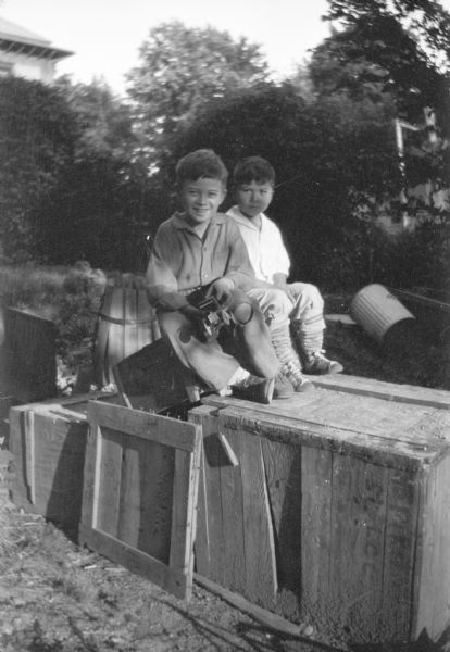 Two unidentified boys sitting on a wooden crate with their feet resting on a larger wooden box. The smiling boy on the left is holding a toy car. There is a wooden barrel with loose staves behind them, and a garbage can is lying on the ground.  