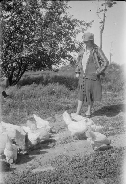 Leonore Judkins Middleton, wearing a striped jacket over her fancy dress, is looking down at a flock of chickens. She is wearing shoes with buckles, and a hat. 