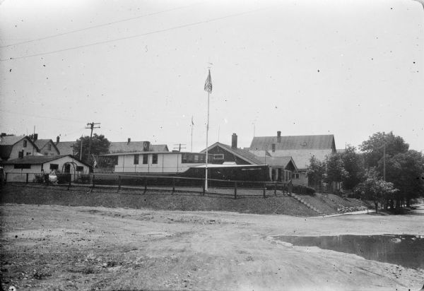 The Edmund B. Gustorf house, built in the 1920's in the shape of a boat, sitting on a low rise at the corner of East Hampshire Street (foreground left) and North Cambridge Avenue. There are portholes in the lower level of the house, and a flag pole is standing in the yard. There is a small building, possibly the garage, of conventional design behind the house. There is a large puddle in the street in the foreground.