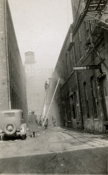 A fireman is climbing a tall ladder supported on the rear wall of a two-story brick building. There are other firemen on the ground and two men observing. There is smoke coming from a second story window, and also filling the air beyond the ladders. A car is parked on the left. In the background is the water tower atop the seven-story Frankfurth Hardware Building (built 1880's, demolished 1980), which faced North Plankinton Avenue.  