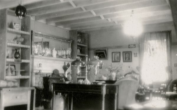 Sunlight and a ceiling fixture provide light for a parlor furnished in an Early American style. A portrait of George Washington is hanging on the far wall. There are exposed joists overhead and a fireplace with mantle on the left. Open shelves flank the fireplace and display glassware and pottery. A double student oil lamp is sitting on a table in the foreground.