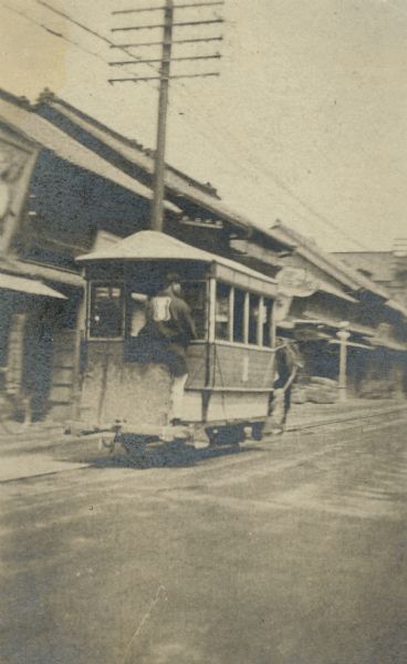 A man is standing on the rear of a small streetcar in Japan. There is a utility pole and several two-story buildings on the left.  