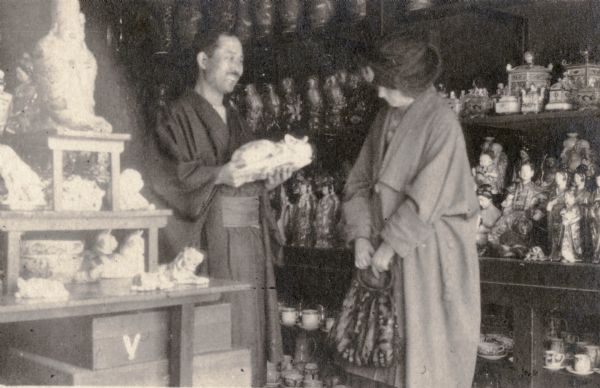 A smiling shopkeeper in traditional dress showing a ceramic cat to Leonore Middleton. There are shelves behind Leonore which are crowded with figurines.