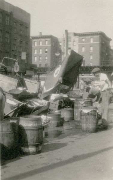 The bed of a truck, barrels and crates are hold fishing and other seafood tended by workers at a fish market. Also pictured are hanging scales and a canvas shade. There is an elevated street or railroad in the background, with multi-story buildings beyond.  