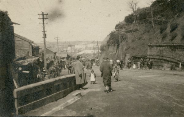 Leonore Middleton, center left, is posing standing along a bridge railing wearing a long winter coat and hat. Other people in traditional Japanese attire are walking in the street. There is a hillside on the right and wooden buildings on the left, and tall utility poles on each side of the street. On the reverse of the print is written: "One of the streets we often take from the Bluff down to the city."  
