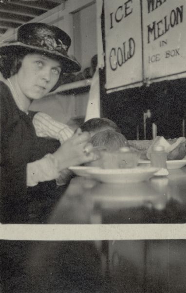 Leonore Middleton sitting at a counter eating a slice of melon.  She is wearing a hat with an artificial flower in the front. A sign behind the counter advertises: "Ice Cold Water Melon in Ice Box." On the reverse of the print is written: "Taken in a little Greek fruit store on Market St. S. F. Sept. 1, 1920."
