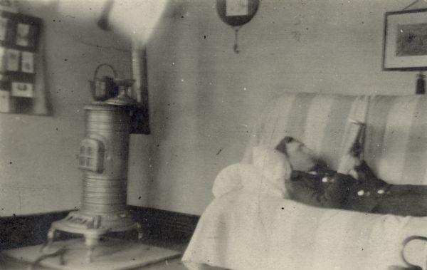 Forest Middleton reading while lying on a sofa in the Middleton's apartment in Japan. The decorative top piece of the stove in the corner has been moved aside to accommodate a large metal kettle. On the reverse of the print is written: "[unreadable] picture but its F's on a Sun. a.m. He still likes to read."