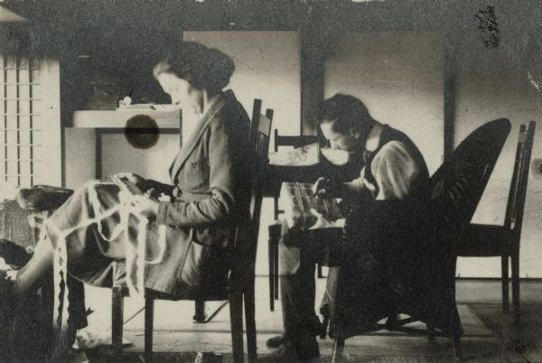 Forest Middleton, right, addressing a package while sitting in a wicker armchair. His wife Leonore, is sitting on a straight back chair with her feet propped on a footstool, wrapping fabric edging on a card. A caption on the reverse of the print states: "Sun. finds us busy marking and doing up things for your export business." The writing most likely addresses Forest's father, William Middleton, who owned a leather goods business in Madison, Wisconsin. He also sold merchandise sent to him by Forest and Leonore from Japan.