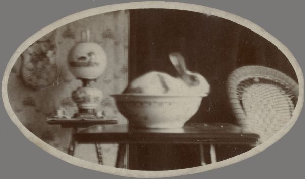 A rabbit, one of the Middleton family pets, resting in a large bowl on a table. There is an oil parlor lamp on a smaller table in the background. The back of a wicker chair is on the right.