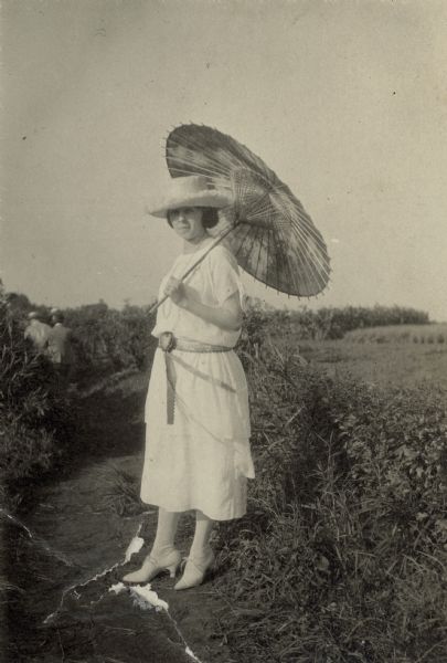 Leonore Middleton posing outdoors, wearing a large hat with a Japanese parasol resting on her left shoulder. She is wearing a light-colored dress with a belt. There are several men in the far left background, facing away from the camera. In the background on the right is a cornfield.