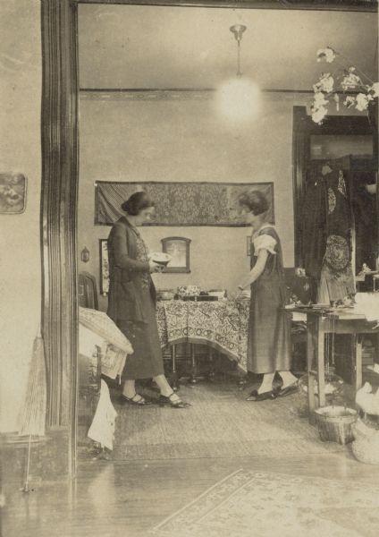Leonore Middleton, left, is holding a bowl, with an unidentified woman  watching on the right. They are standing in front of a table which is holding other bowls and objects. There are printed textiles on the table, with more hanging on the wall and on the open door of a closet.  On the floor are baskets and two duck figurines.