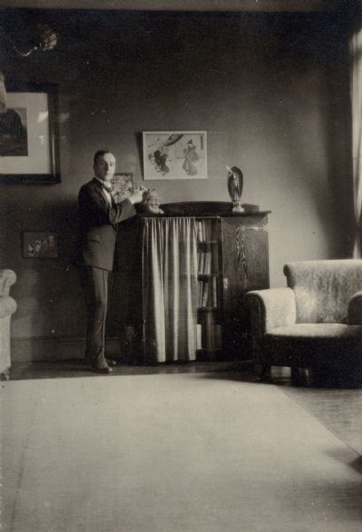 Forest Middleton standing beside a bookcase holding a pipe. He is wearing a suit and tie. There are Japanese figurines on the top of the bookcase and a Japanese print hanging on the wall. There are upholstered armchairs on each side of the bookcase, and a carpet on the floor.