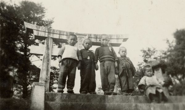 Four young boys standing, with a fifth child sitting, on the top step in front of a Japanese torii, or gate. There is a stone lantern at far right. On the reverse of the print is written: "Shrine at Shirakibaru."