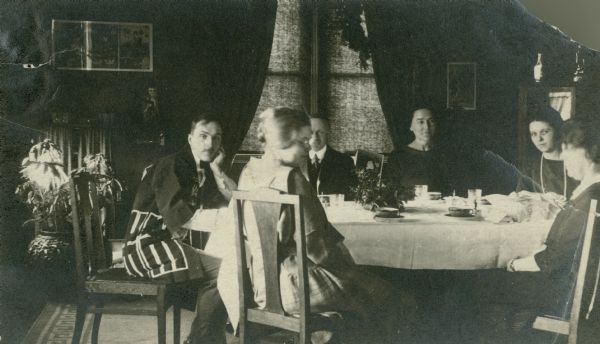 Four women and two men are sitting at a dining table. Forest Middleton, on the far left, is wearing a Japanese-style jacket. His wife, Leonore, is second from right and is wearing a long bead necklace. There is a small floral arrangement on the table and broad, flat cups in saucers. On the reverse of the print is written: "The last course of our Christmas dinner. Very good of the cups & Forest."