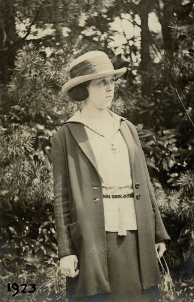 Leonore Middleton posing out of doors for a three-quarter length standing portrait. She is wearing a blouse and skirt with a light coat and a summer hat. There is shrubbery in the background. On the reverse of the print is written: "Taken by a Waseda University student June 14 '23. Tokyo, Japan."