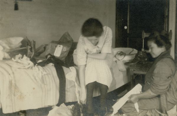 Leonore Middleton leaning forward as she is sitting on a bed trying on a pair of shoes. An unidentified Japanese woman in traditional dress is sitting on the floor, right. The bed is strewn with clothes and packing materials. On the reverse of the print is written: "3 prs. of shoes & 1 pr. rubbers arrive from Am[erica]. They all fit, fine."