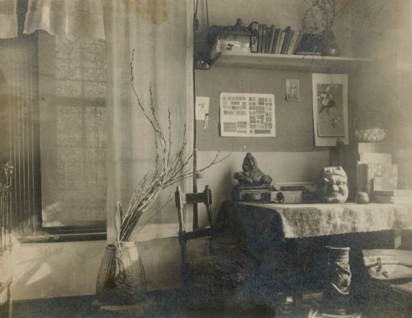 A view of an interior corner of the house occupied by Forest and Leonore Middleton. There are light draperies and a shade over a large window. A bouquet of pussy willow branches extend from a basket on the floor. In the corner is a table with a print cloth on which figurines and books rest. A page of mounted stamps is hanging on the wall near a Japanese print. There is a bookshelf on the wall.  